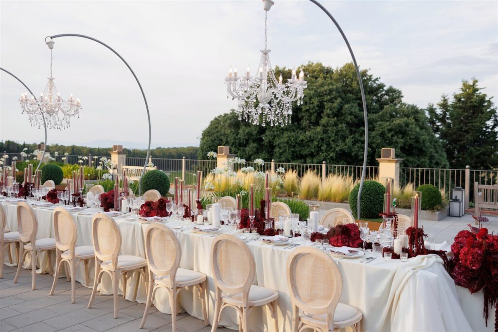 An Unforgettable Anniversary at Castello del Nero: A Three-Day Celebration in Tuscany 36 Elegant outdoor wedding reception table setting with chandeliers and red floral decor. Anniversary Castello del Nero - Italian Wedding Designer