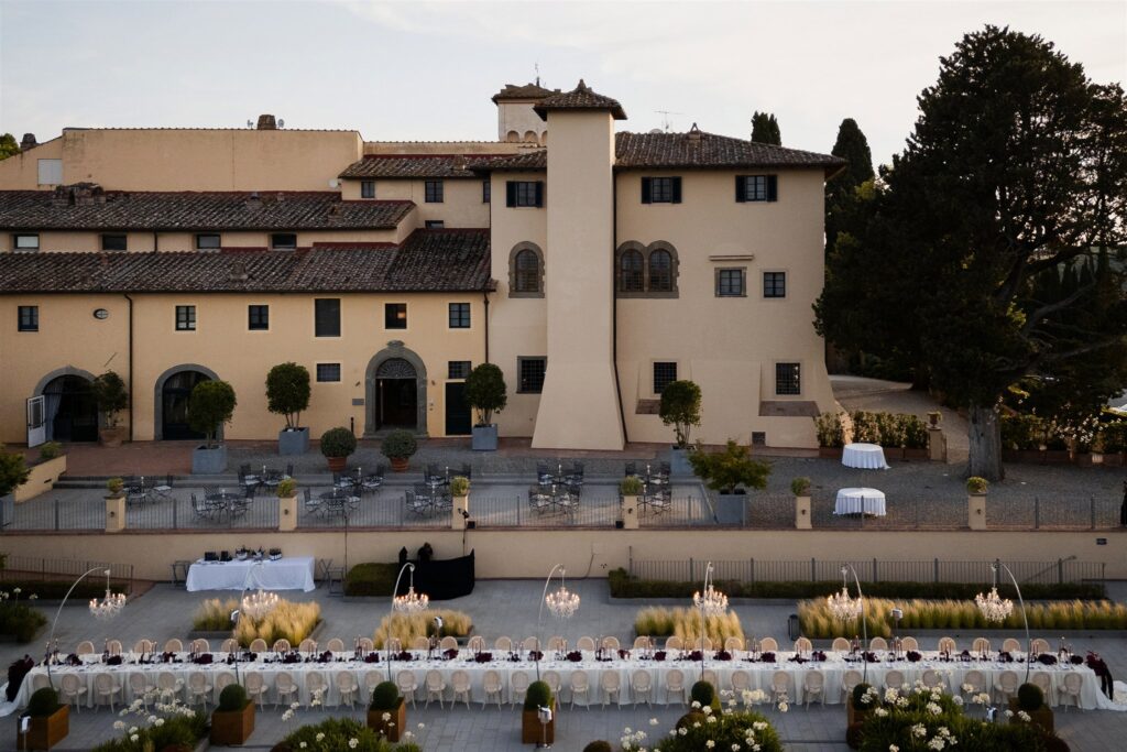 An Unforgettable Anniversary at Castello del Nero: A Three-Day Celebration in Tuscany 39 Elegant outdoor wedding reception setup with long tables and chandeliers in front of a Tuscan. Anniversary Castello del Nero - Italian Wedding Designer villa.
