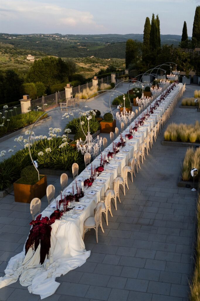 An Unforgettable Anniversary at Castello del Nero: A Three-Day Celebration in Tuscany 40 Elegant long table setting with red floral arrangements and chandeliers outdoors. Anniversary Castello del Nero - Italian Wedding Designer