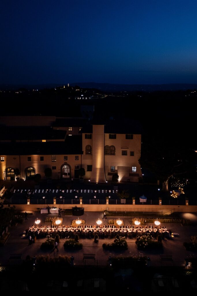 An Unforgettable Anniversary at Castello del Nero: A Three-Day Celebration in Tuscany 48 Elegant outdoor long table dinner setup at dusk with illuminated building. Anniversary at Castello del Nero - Italian Wedding Designer