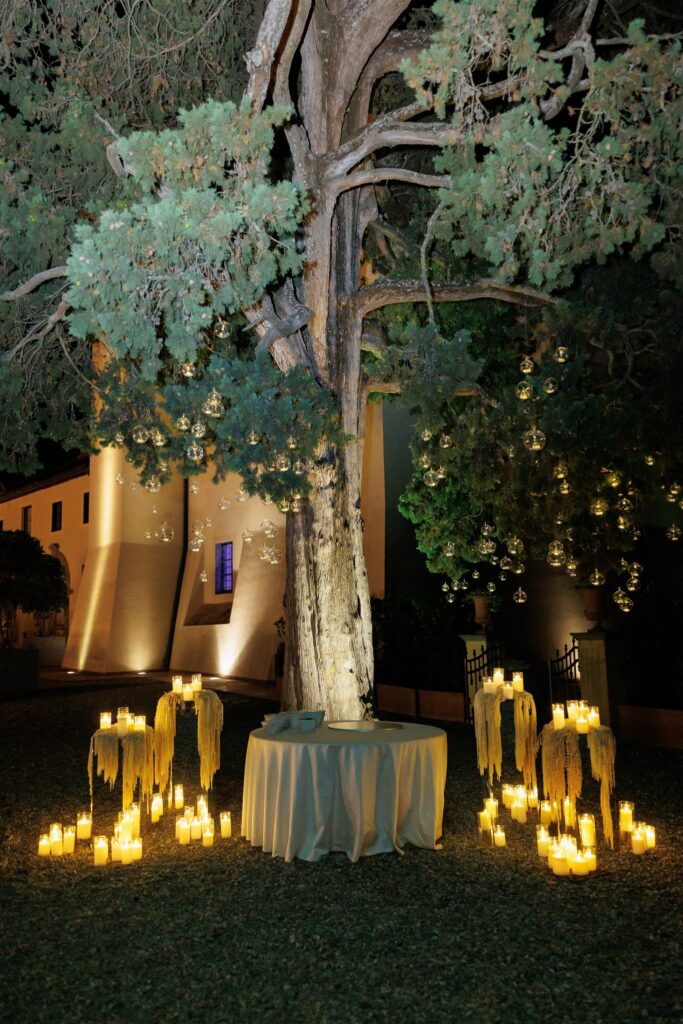 An Unforgettable Anniversary at Castello del Nero: A Three-Day Celebration in Tuscany 47 Magical outdoor wedding setup with glowing lanterns & candles under a large tree at night. Anniversary Castello del Nero - Italian Wedding Designer