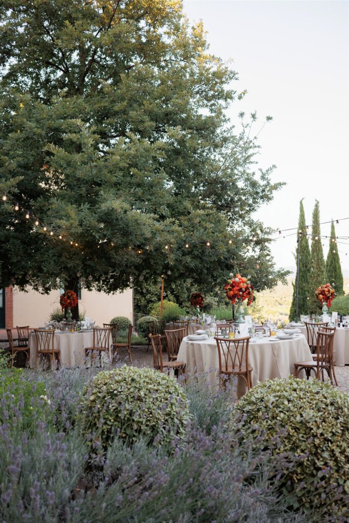 An Unforgettable Anniversary at Castello del Nero: A Three-Day Celebration in Tuscany 16 Elegant outdoor wedding reception tables set with floral centerpieces under string lights in a garden. Anniversary Castello del Nero - Italian Wedding Designer