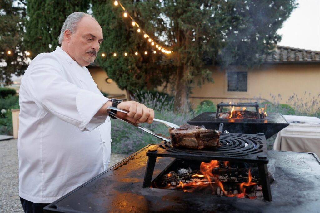An Unforgettable Anniversary at Castello del Nero: A Three-Day Celebration in Tuscany 17 Chef grilling a large steak over open flames outdoors at dusk. Anniversary Castello del Nero - Italian Wedding Designer