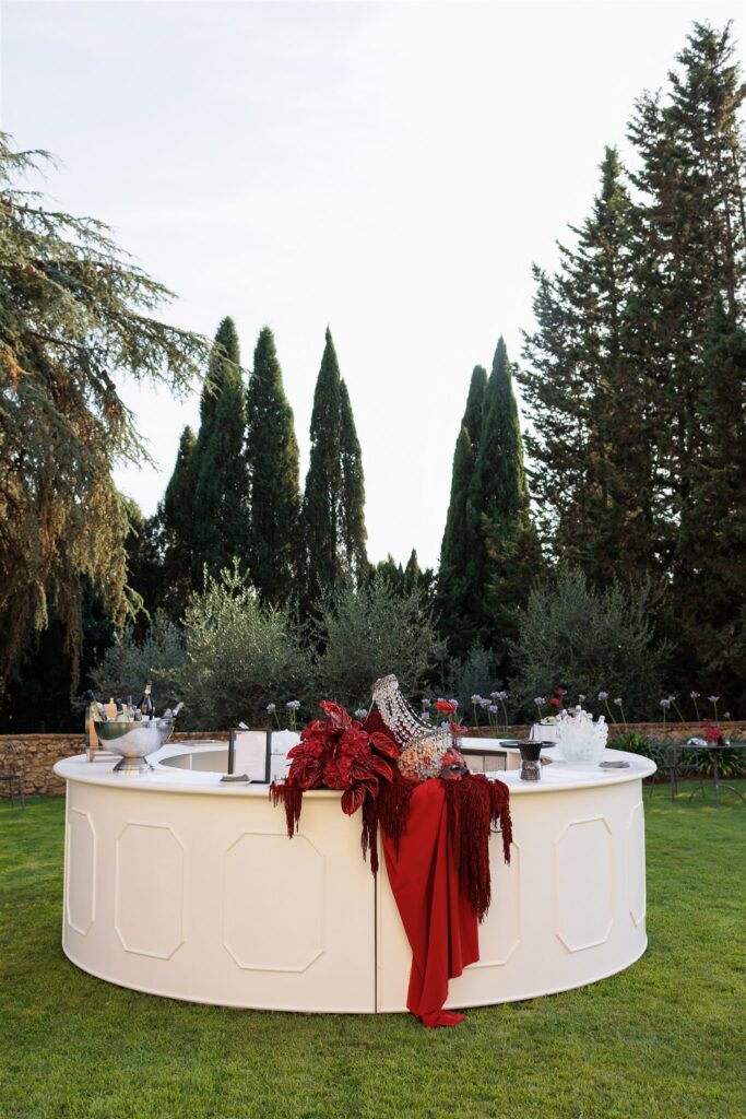 An Unforgettable Anniversary at Castello del Nero: A Three-Day Celebration in Tuscany 25 Elegant white bar set up outdoors with red floral decor and champagne. Anniversary Castello del Nero - Italian Wedding Designer