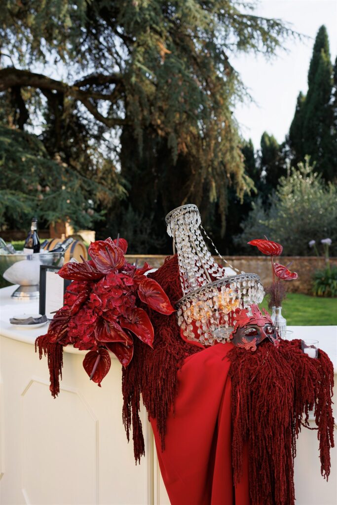 An Unforgettable Anniversary at Castello del Nero: A Three-Day Celebration in Tuscany 26 Masquerade bar setup with red flowers, crystal chandelier, and masks outdoors. Anniversary Castello del Nero - Italian Wedding Designer