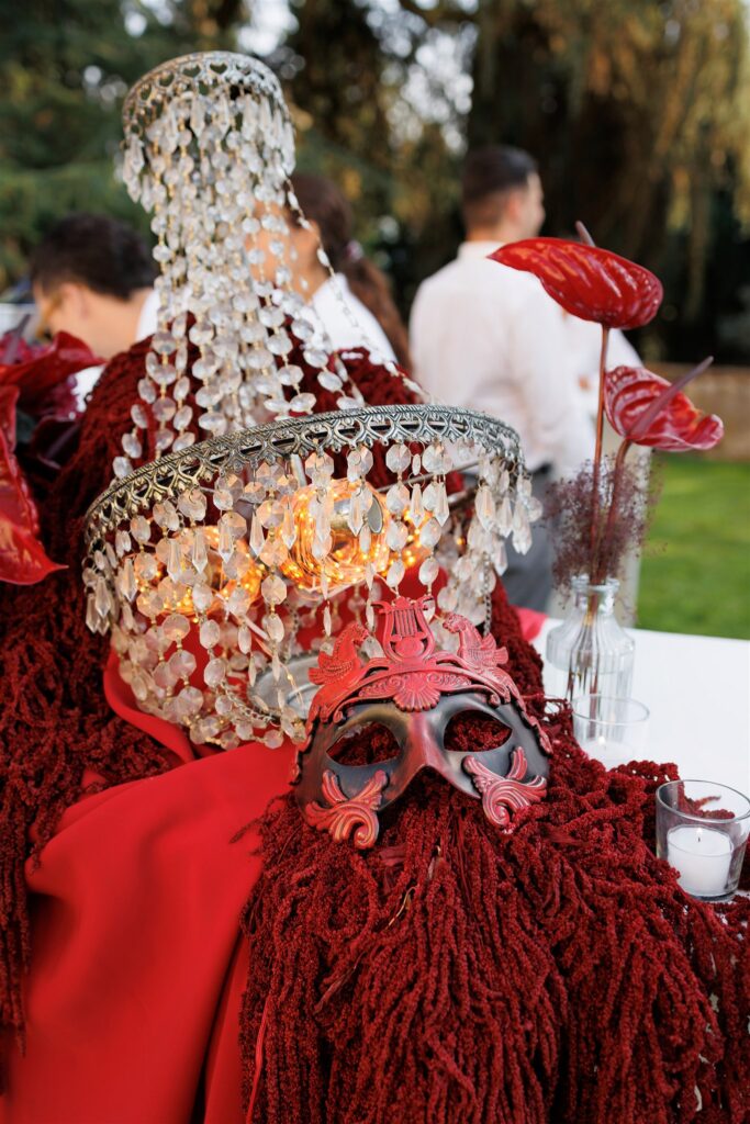 An Unforgettable Anniversary at Castello del Nero: A Three-Day Celebration in Tuscany 27 Elegant masquerade mask and crystal chandelier centerpiece with red accents outdoors. Anniversary Castello del Nero - Italian Wedding Designer