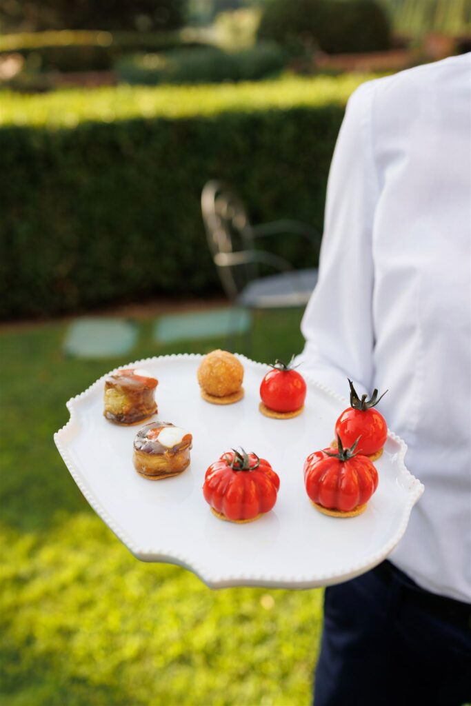An Unforgettable Anniversary at Castello del Nero: A Three-Day Celebration in Tuscany 28 Caterer holding a white plate with assorted appetizers, including tomato-shaped bites and small pastries. Anniversary Castello del Nero - Italian Wedding Designer