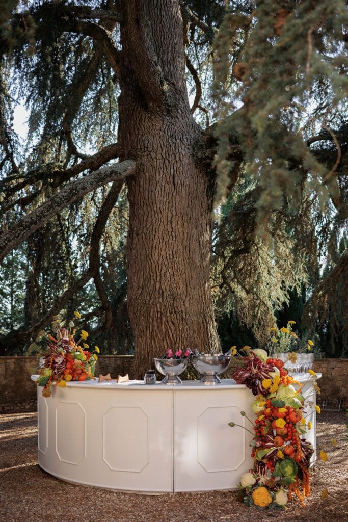 An Unforgettable Anniversary at Castello del Nero: A Three-Day Celebration in Tuscany 4 White curved bar decorated with autumn flowers and produce under a large tree.